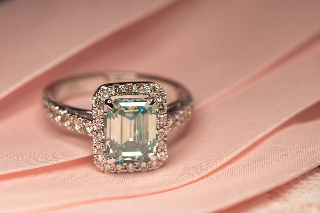 a silver engagement ring featuring a radiant cut center stone and halo setting on a piece of pink fabric