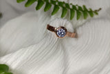 A close-up of a bezel set rose gold engagement ring with a round cut diamond wrapped in white fabric.