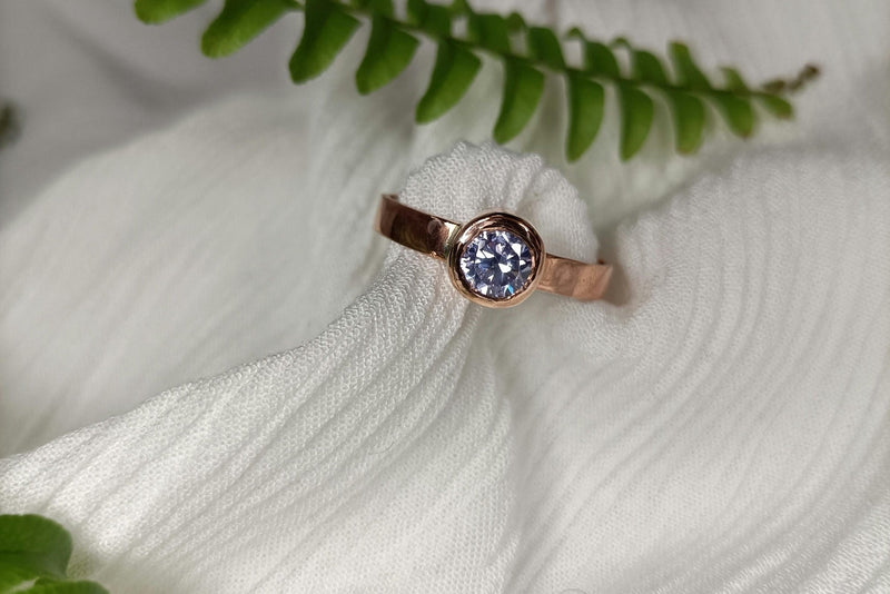 A close-up of a bezel set rose gold engagement ring with a round cut diamond wrapped in white fabric.