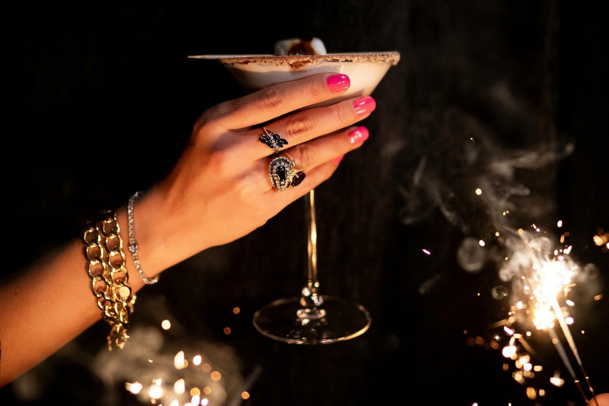 A woman toasting with a smores cocktail while wearing diamond and sapphire statement rings, a couple chunky gold chain bracelets, and a diamond tennis bracelet