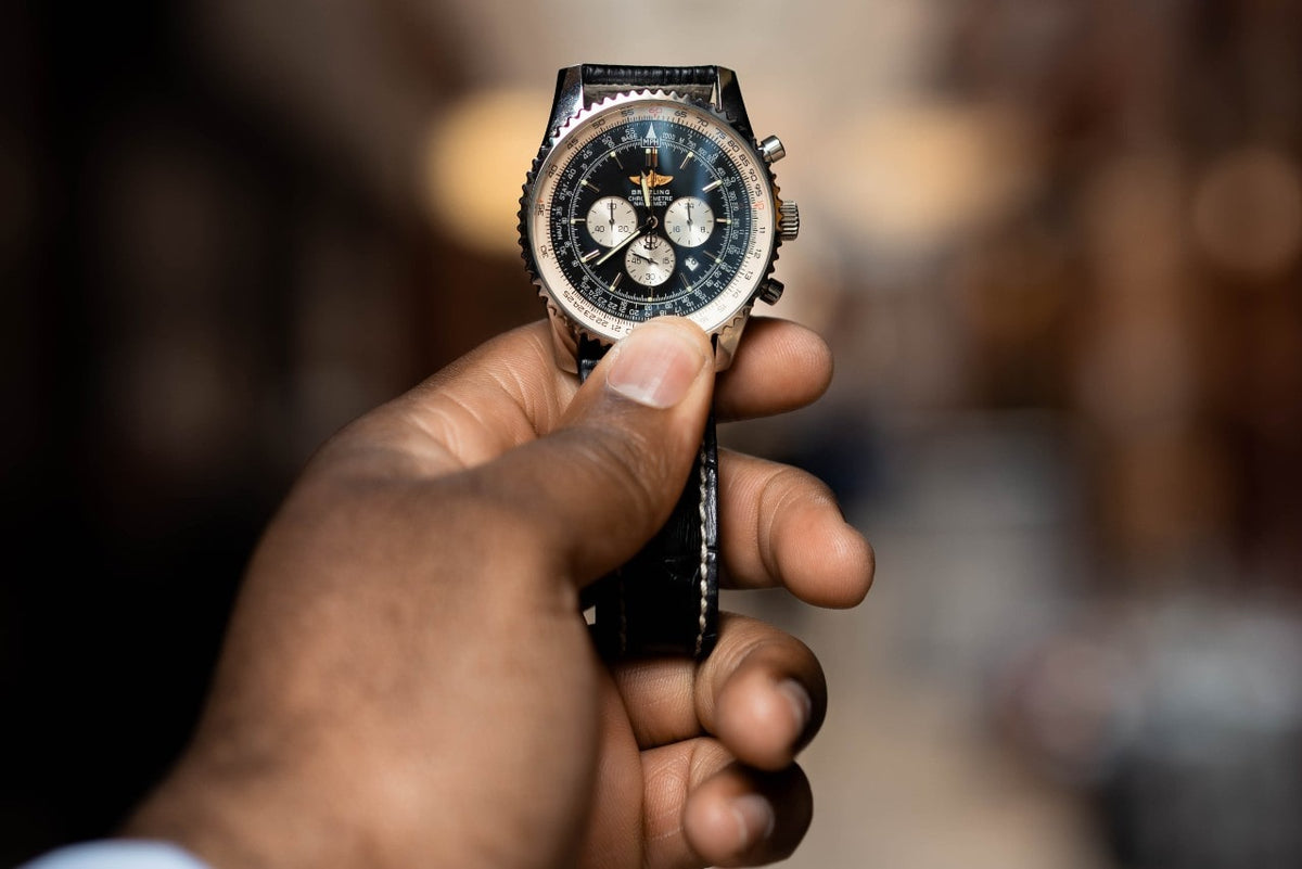 A hand holding a black leather Breitling wristwatch with several complications against a blurry background