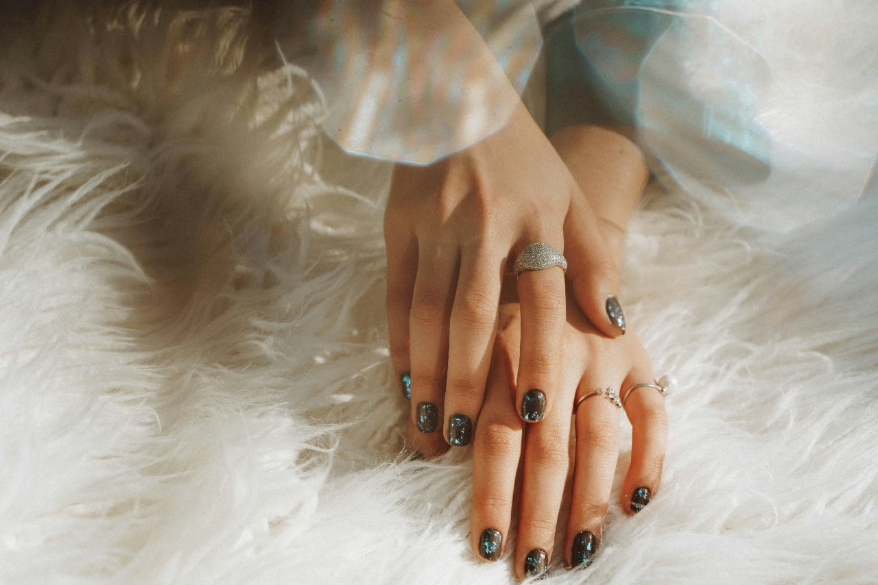 A close up of a hands with brown and blue manicure resting on a white carpet, showcasing various fashion rings