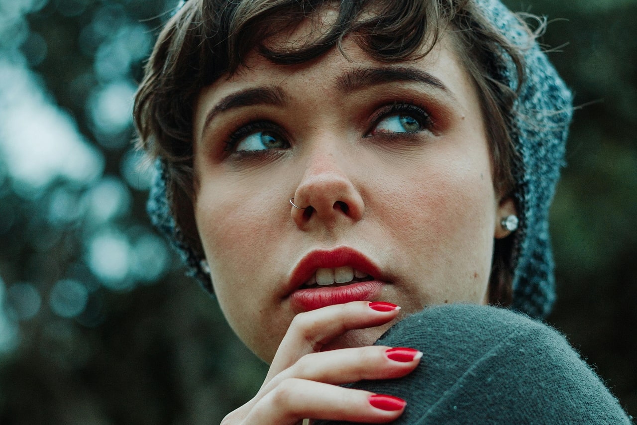 A woman with short hair and beanie gazing away, demonstrating her red nails and diamond studs.