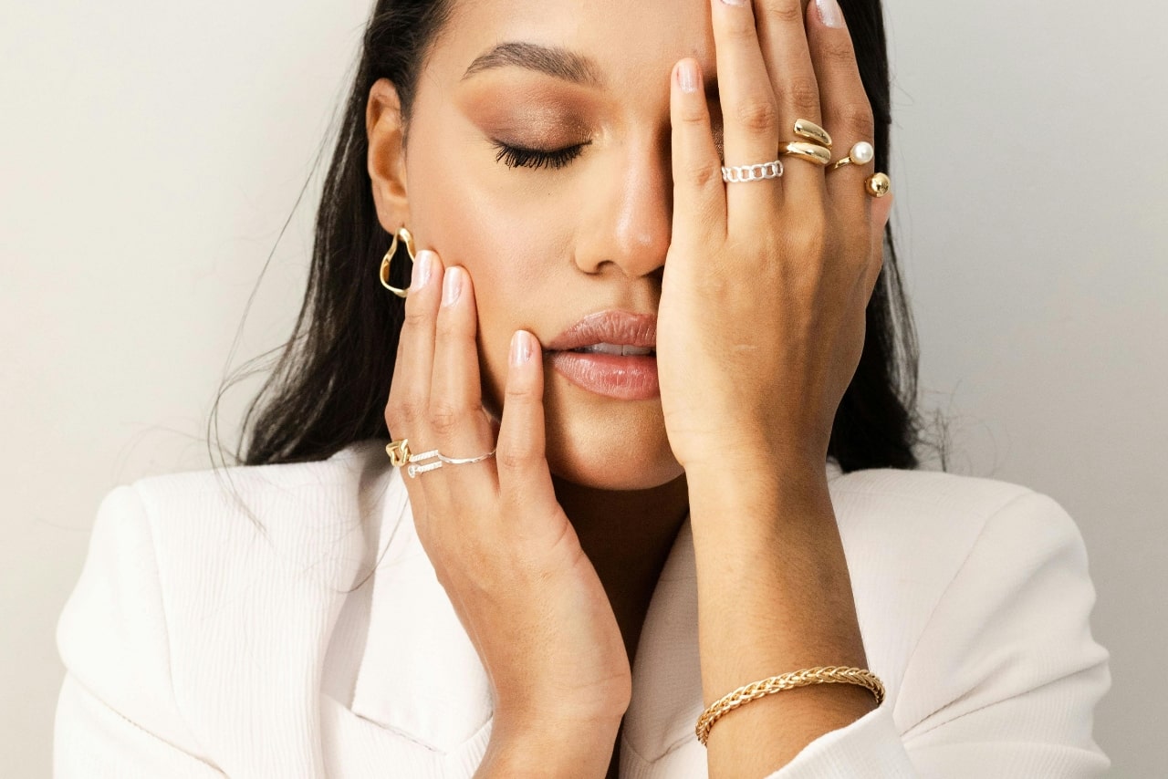 A woman in white blazer with her eyes closed, covering her face with a hand adorned in gold and silver rings.