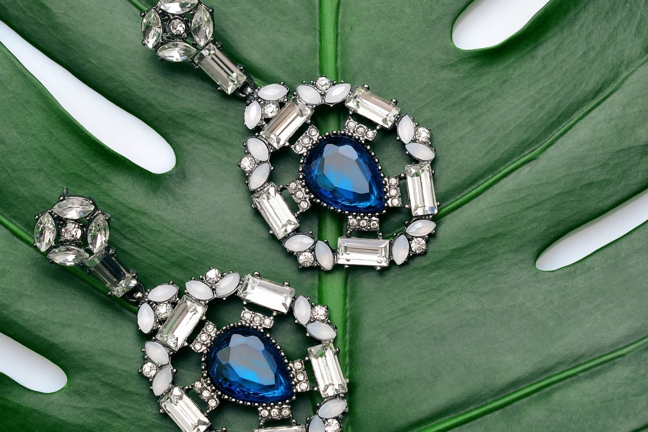 A pair of sparkly diamond earrings with sapphire stones at the center, laying on a leaf.
