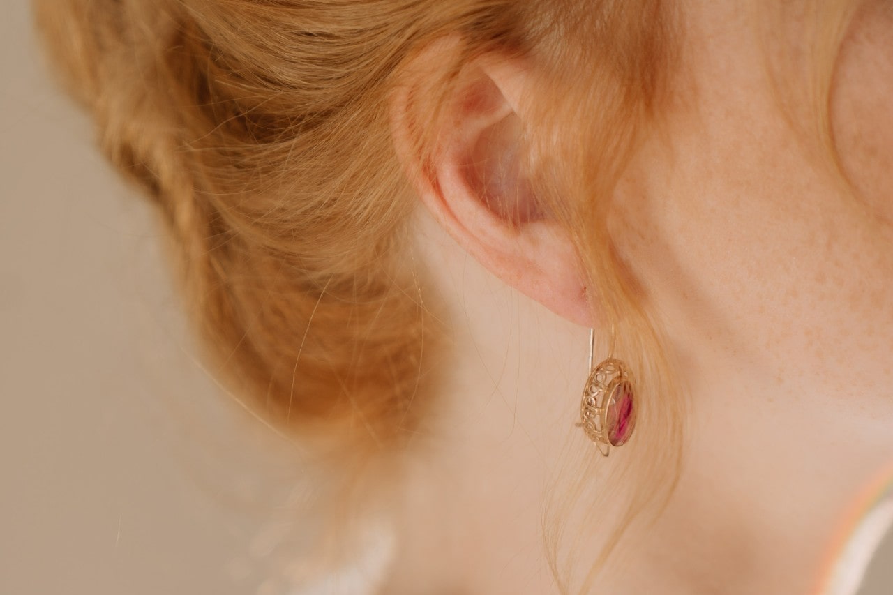 A close up of a woman with bright ginger hair wearing a golden dangly earring with a ruby gemstone.