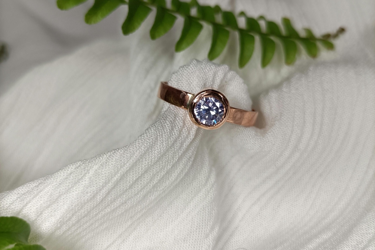 A close-up of a bezel set rose gold engagement ring with a round cut diamond wrapped in white fabric.