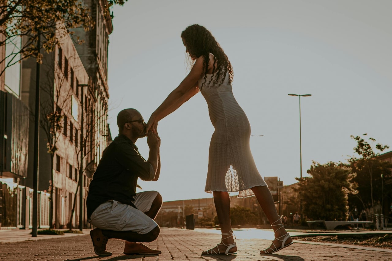 Man kneeling and kissing a woman's hand with an engagement ring during a golden hour city proposal.
