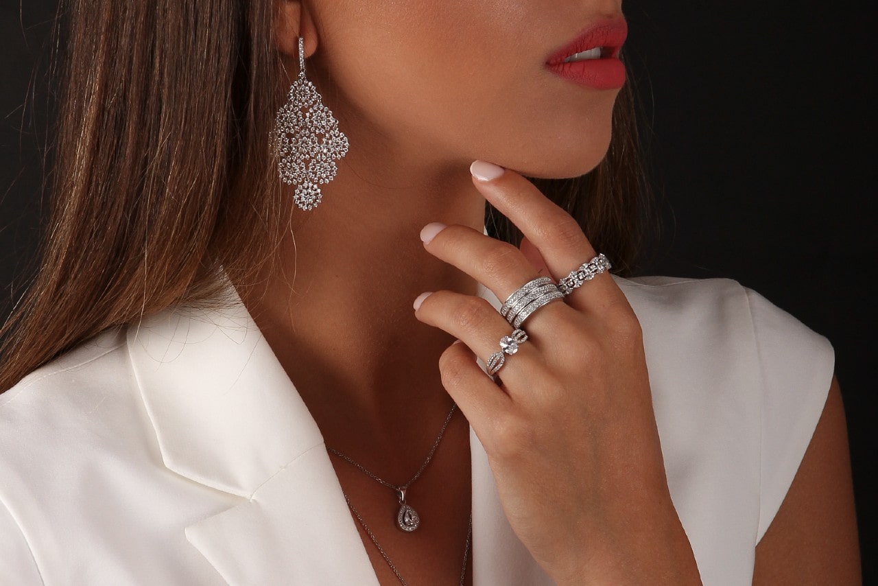 A close up of a woman in white vest, wearing a white gold diamond drop necklace, ornate drop earrings and three fashion rings.