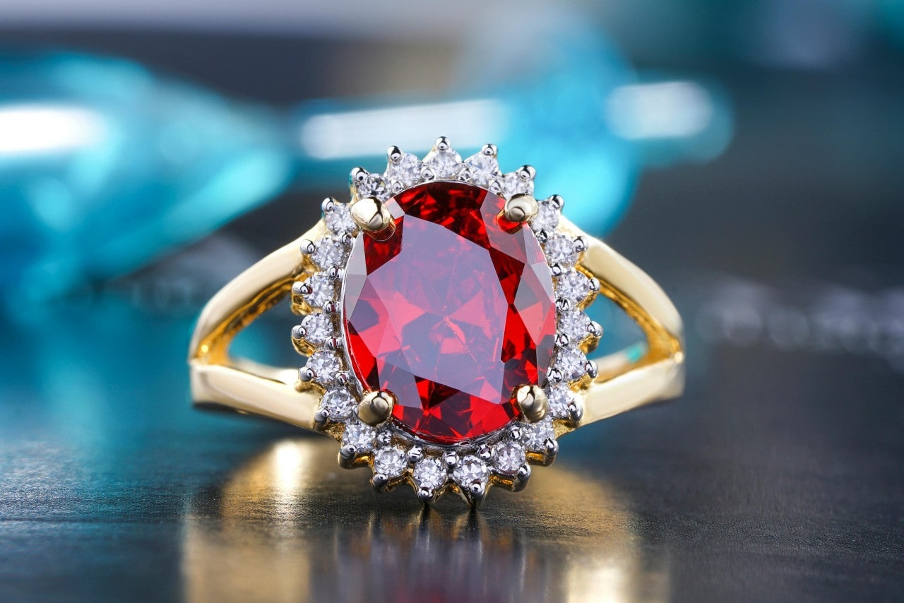 A yellow gold halo engagement ring with oval ruby as a center stone on a grey matte surface.