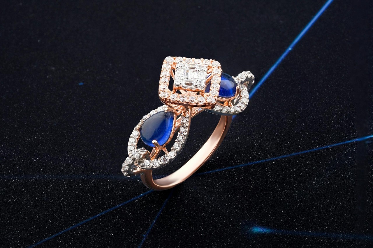 A rose gold ring featuring a square diamond halo centerpiece flanked by two pear-shaped blue gemstones, all accented with pavé diamonds, is displayed against a dark, starry background.