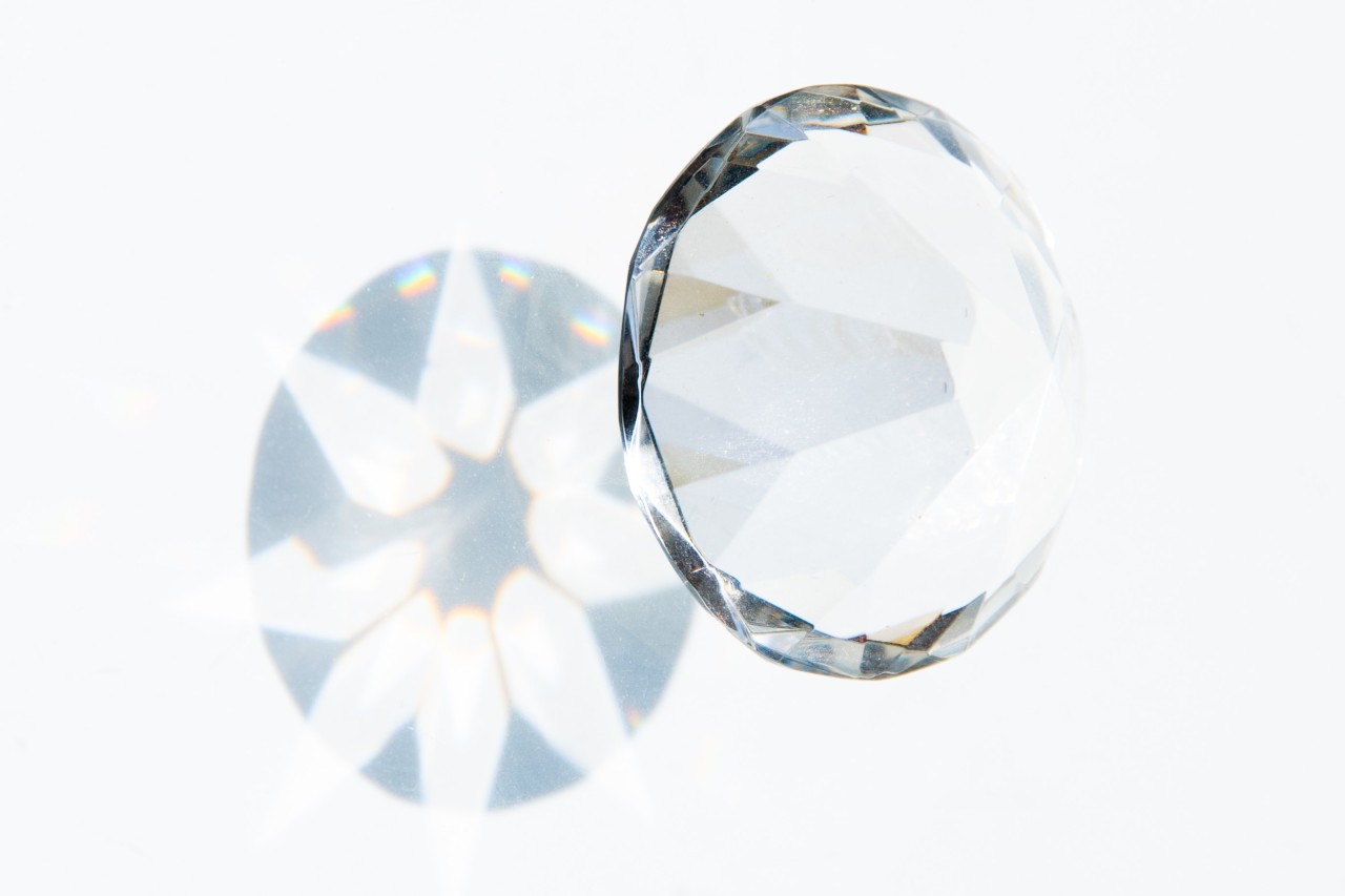 A close-up of a diamond against a white wall, the diamond's reflection shining behind it.