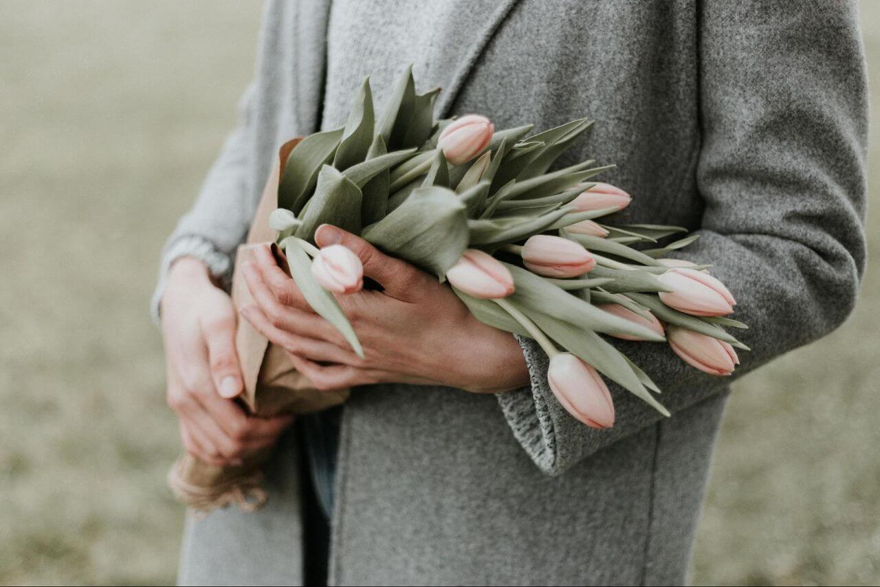 A man in a long gray coat holding light pink tulips in an open field.