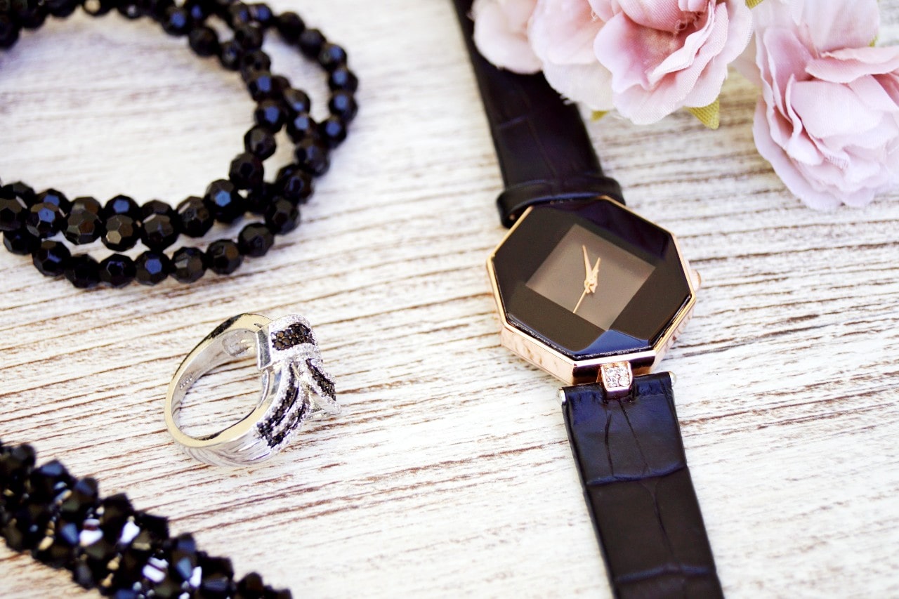 A close-up of a distinctive luxury watch, an elegant fashion ring with black gems, and a black beaded necklace displayed on a pale wooden surface next to pink peonies.