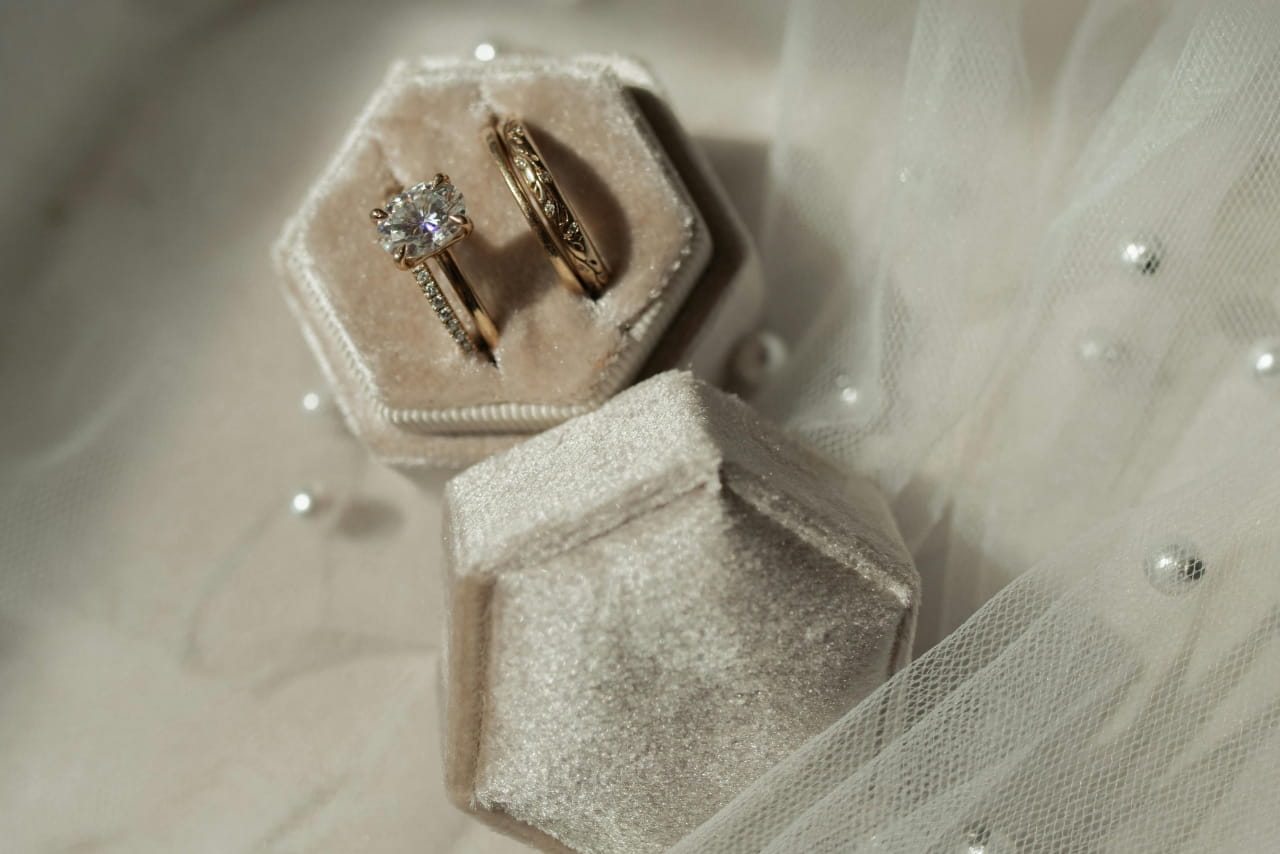 A close-up of an ivory ring box with an engagement ring and a yellow gold wedding band, laying on top of tulle fabric with pearl.