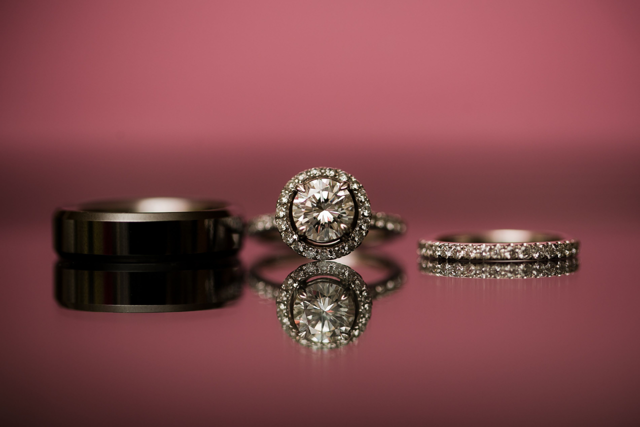 A close-up of a men’s wedding band, a halo engagement ring, and a women’s diamond wedding band on a reflective surface.