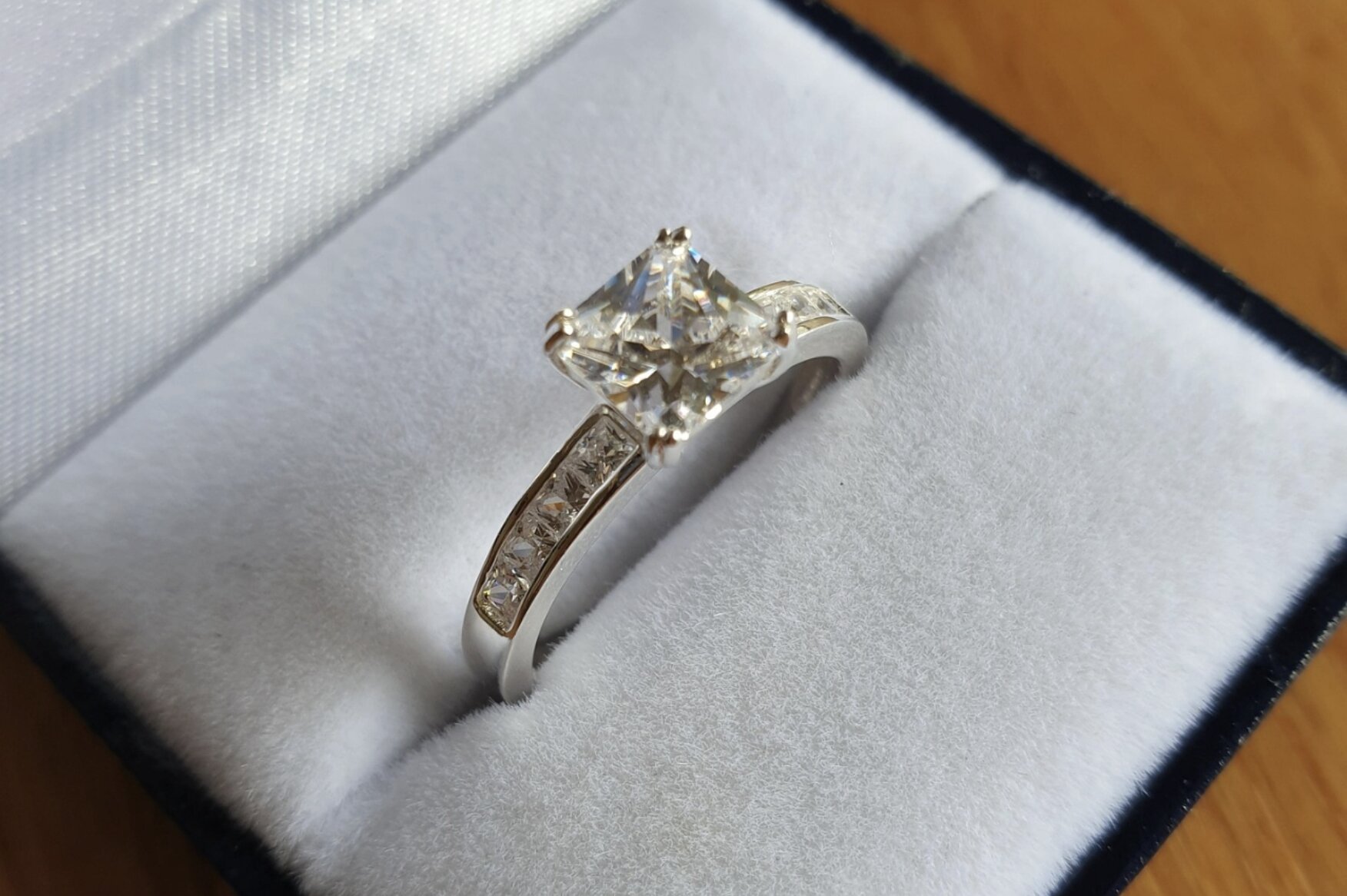 A close-up of a princess cut side stone ring in a light gray ring box on a wooden table.