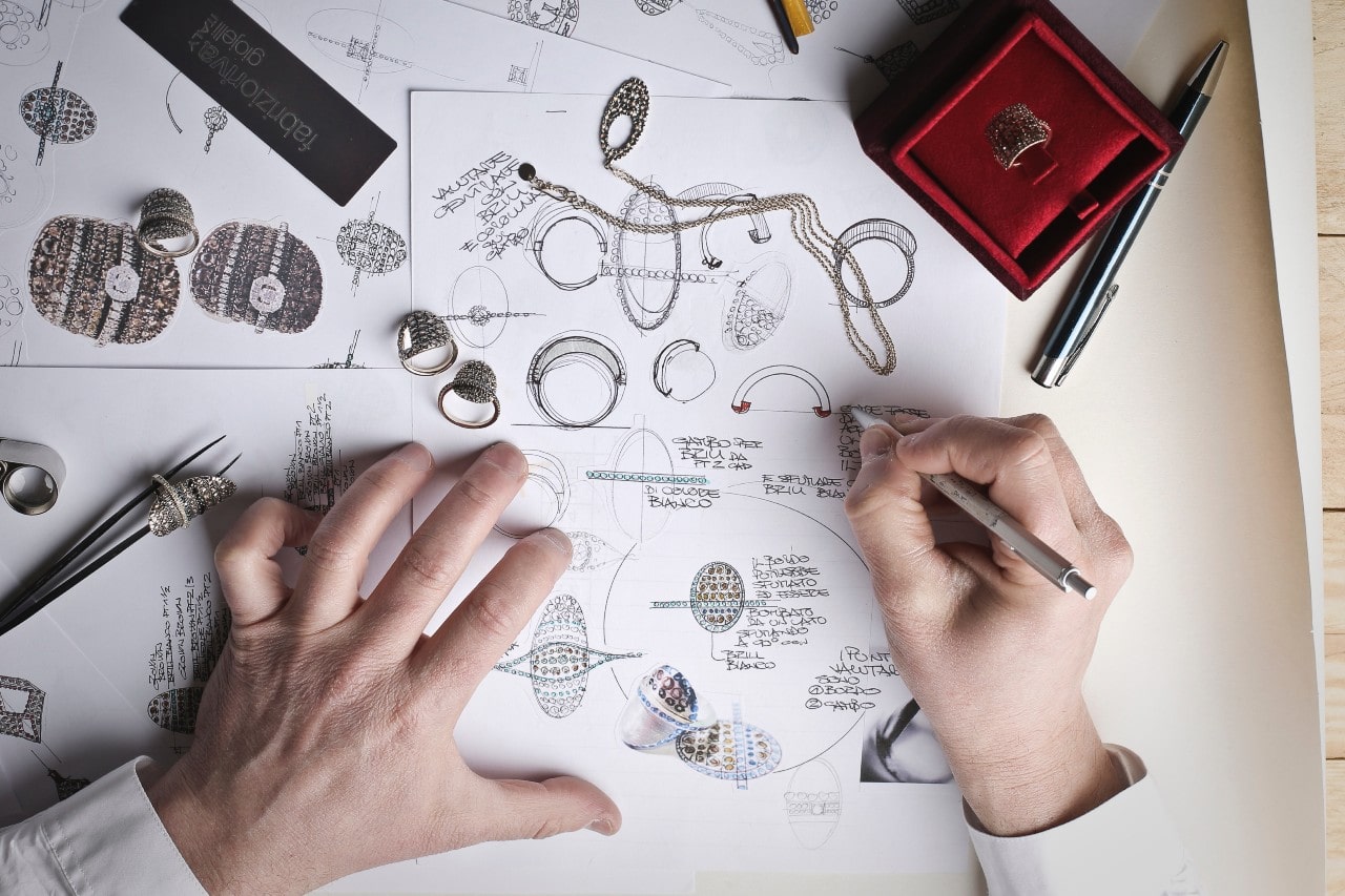 A top-down view of jewelry designer’s hands as they draw designs on a large piece of paper.