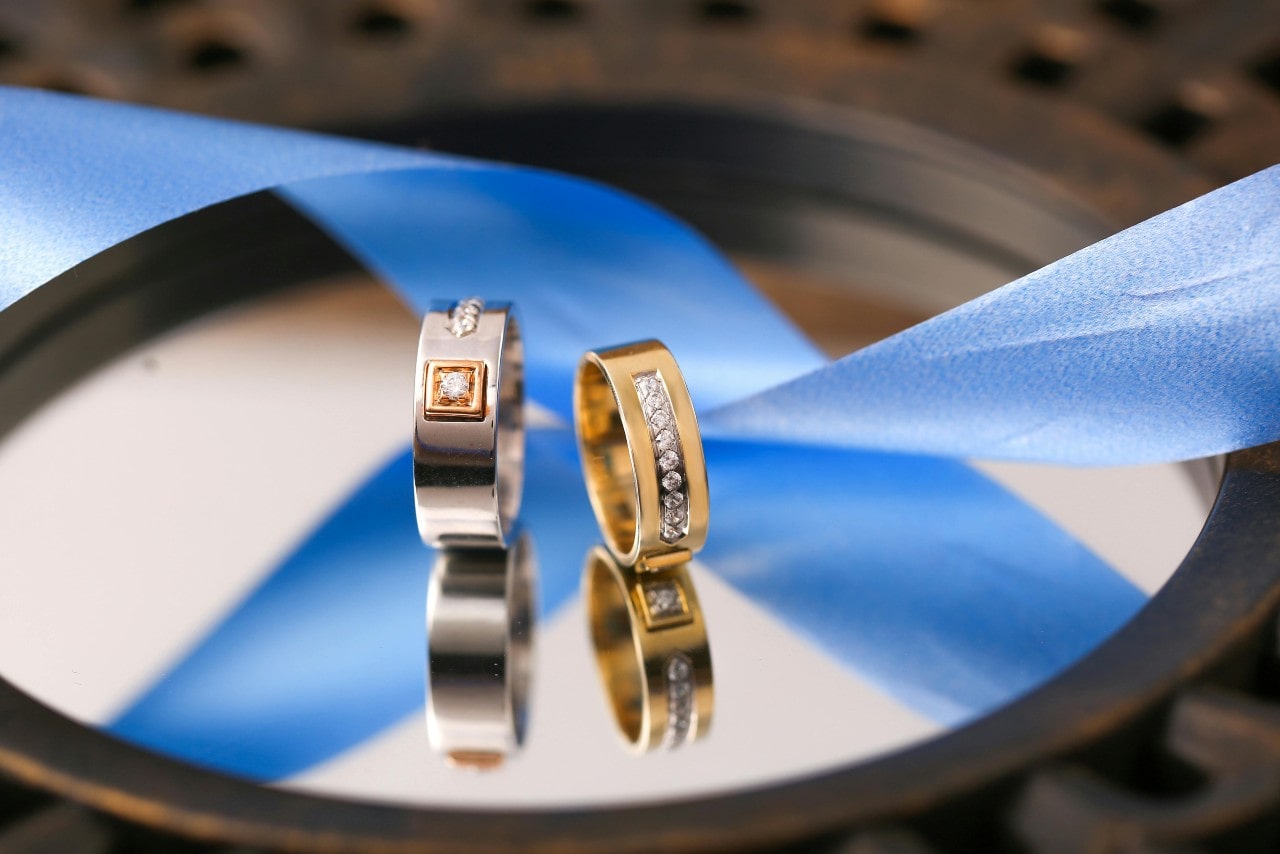 A close-up of two mixed metal diamond wedding bands displayed atop a mirror in front of a blue ribbon.