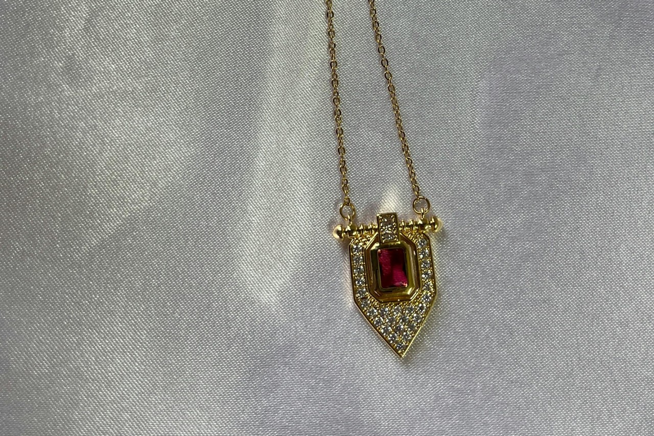A close-up of a yellow gold pendant necklace with a ruby center stone and diamond accents displayed atop textured grey fabric.
