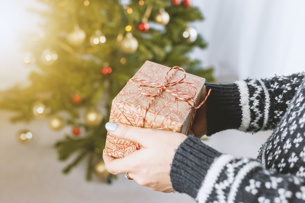 A person in the knitted white and gray sweater holds a Christmas gift with the blurred Christmas tree in the background