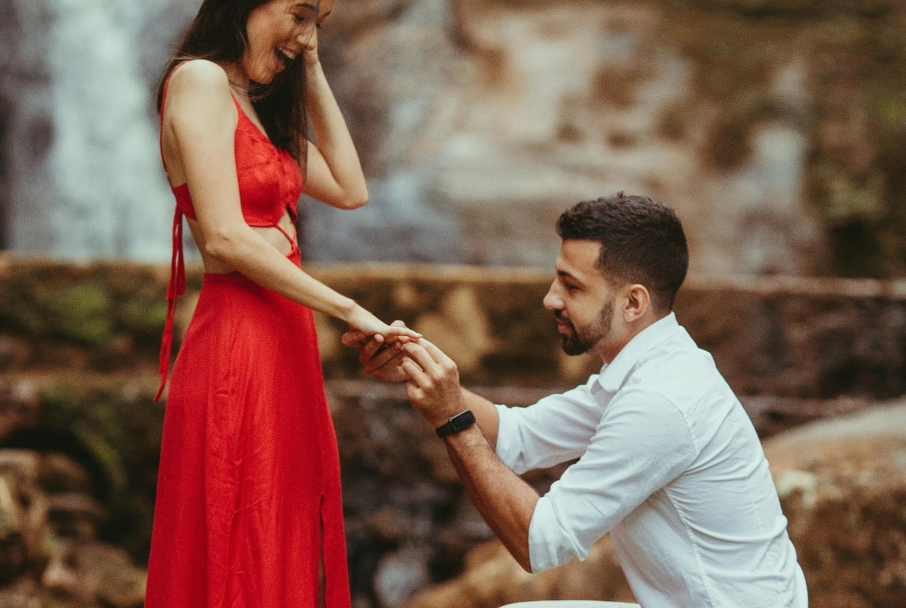 A man kneels to propose to a woman in a vibrant red dress, holding her hand in a lush outdoor setting.
