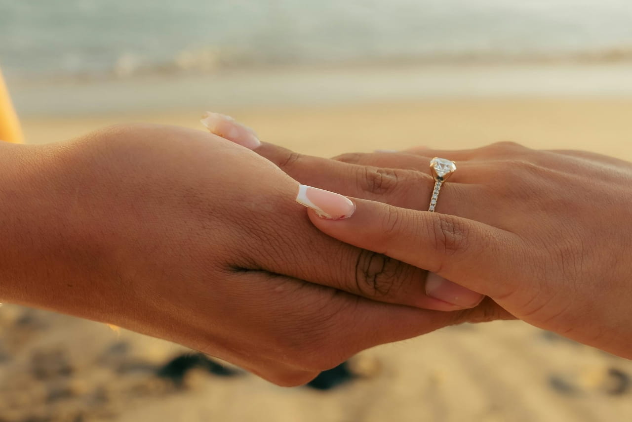 Two hands in front of the beach, featuring a diamond side stone engagement ring on a woman’s hand.