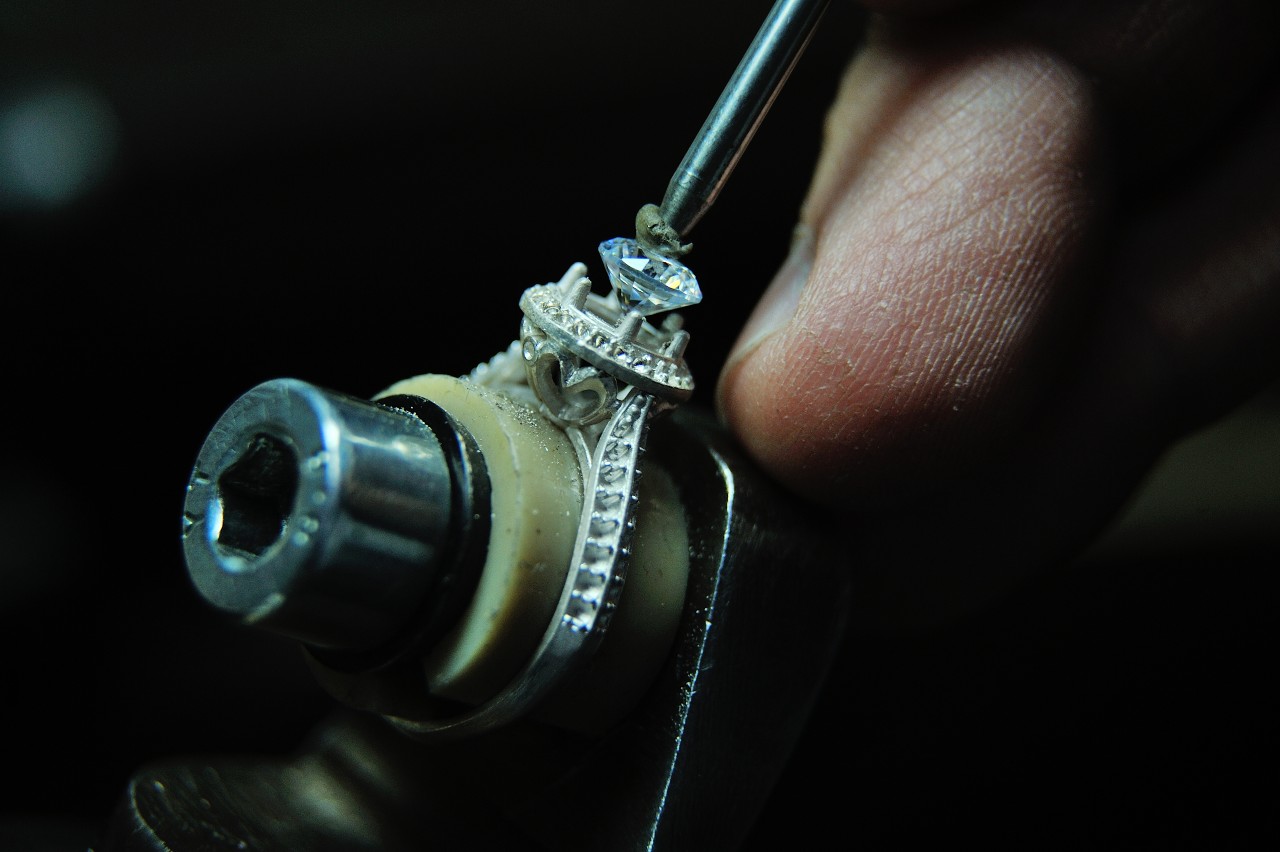 A close-up of a craftsman using a tool to secure a diamond in a halo white gold engagement ring.