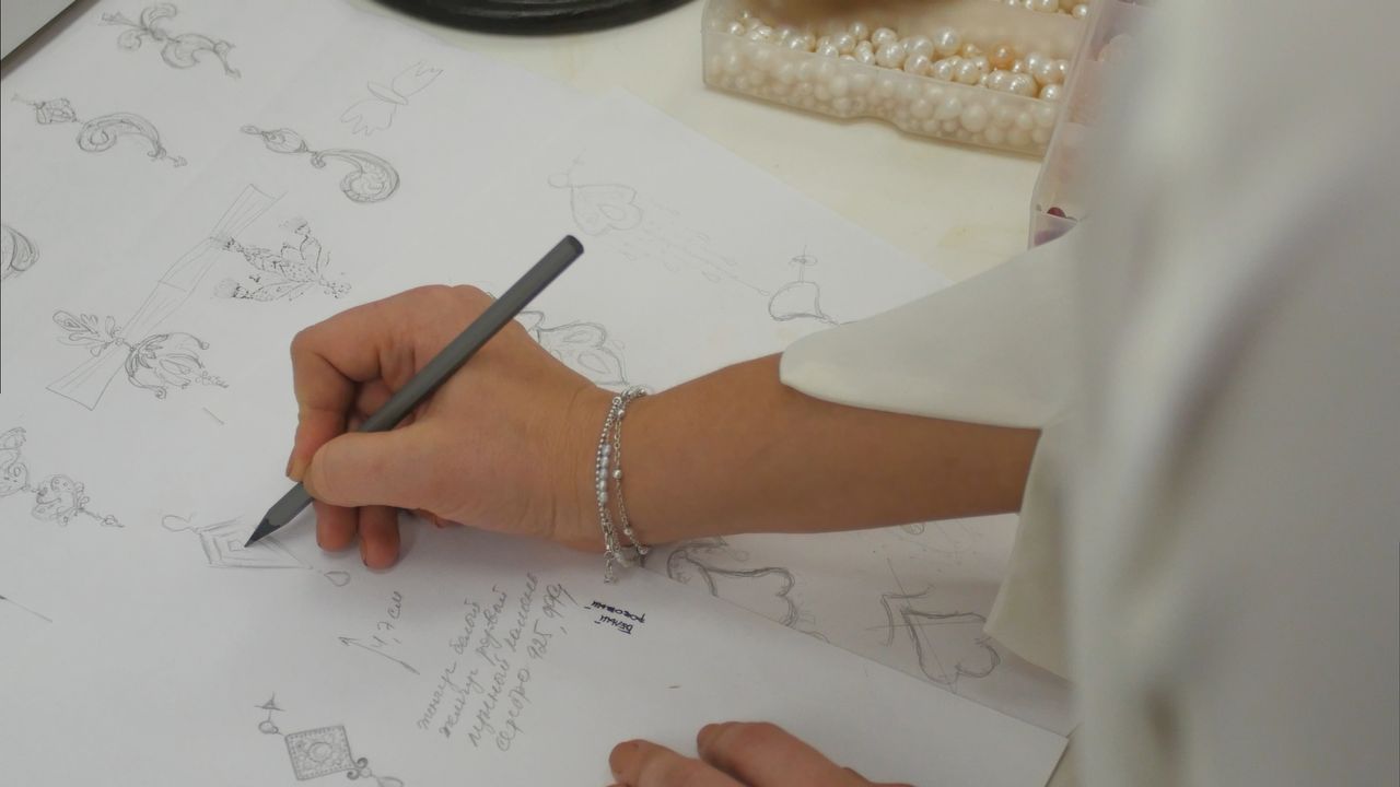 A designer sketches jewelry designs on paper beside a tray of pearls.