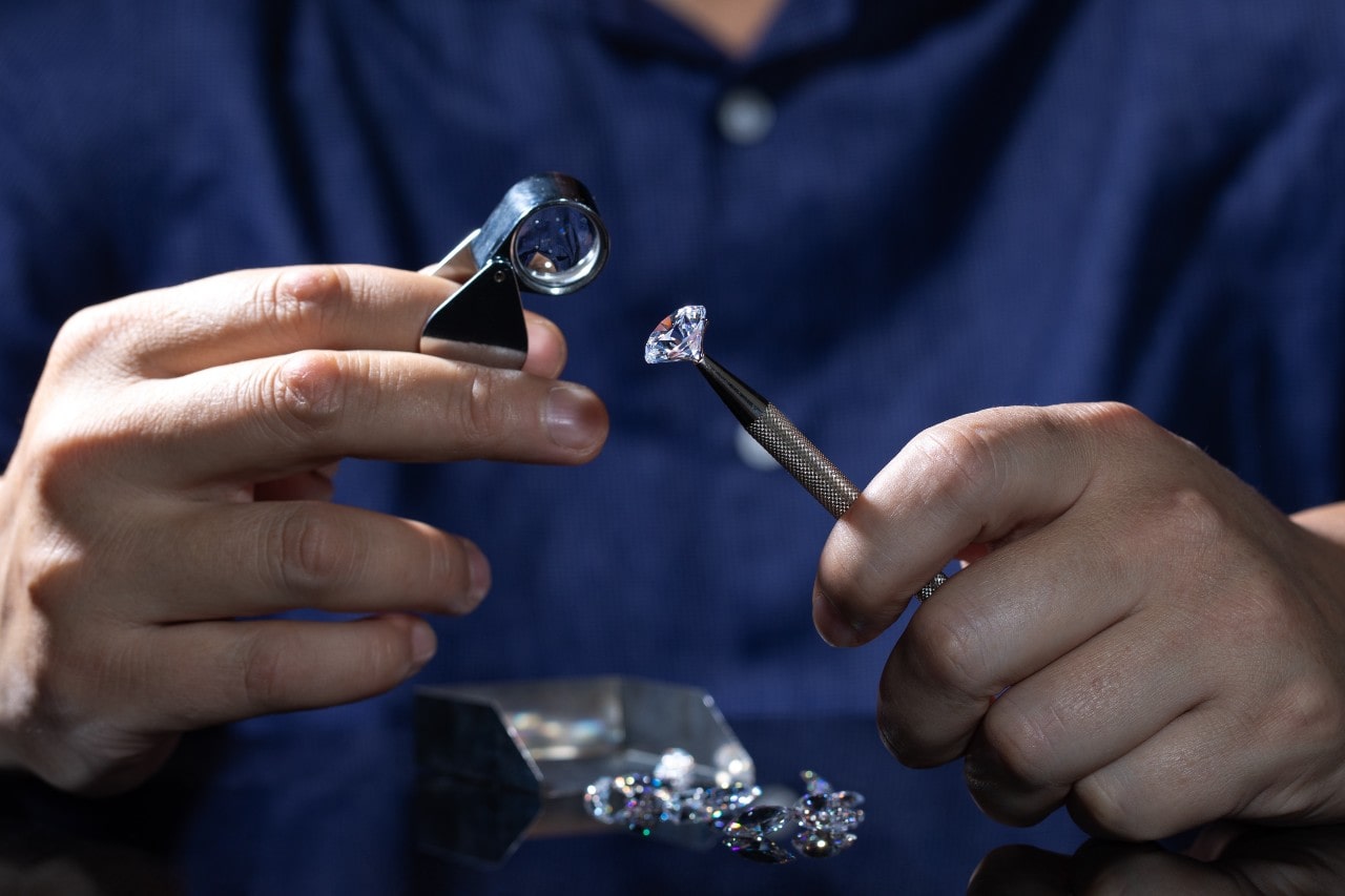 Close-up of a jeweler wearing a dark blue shirt, examining a sparkling diamond with a loupe in one hand while holding it with tweezers in the other.