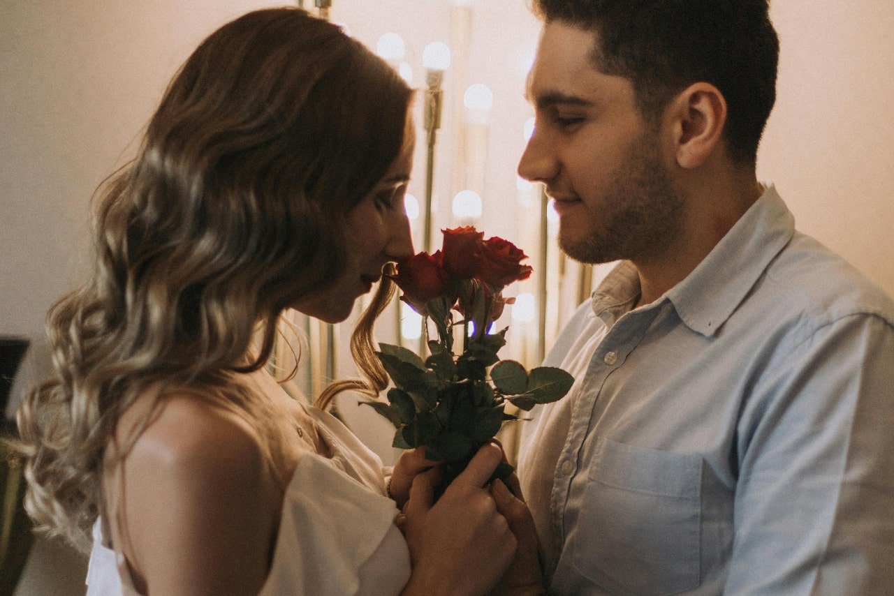 The woman closes her eyes, inhaling the smell of red roses, while the man gazes at her, surrounded by soft, warm lighting.