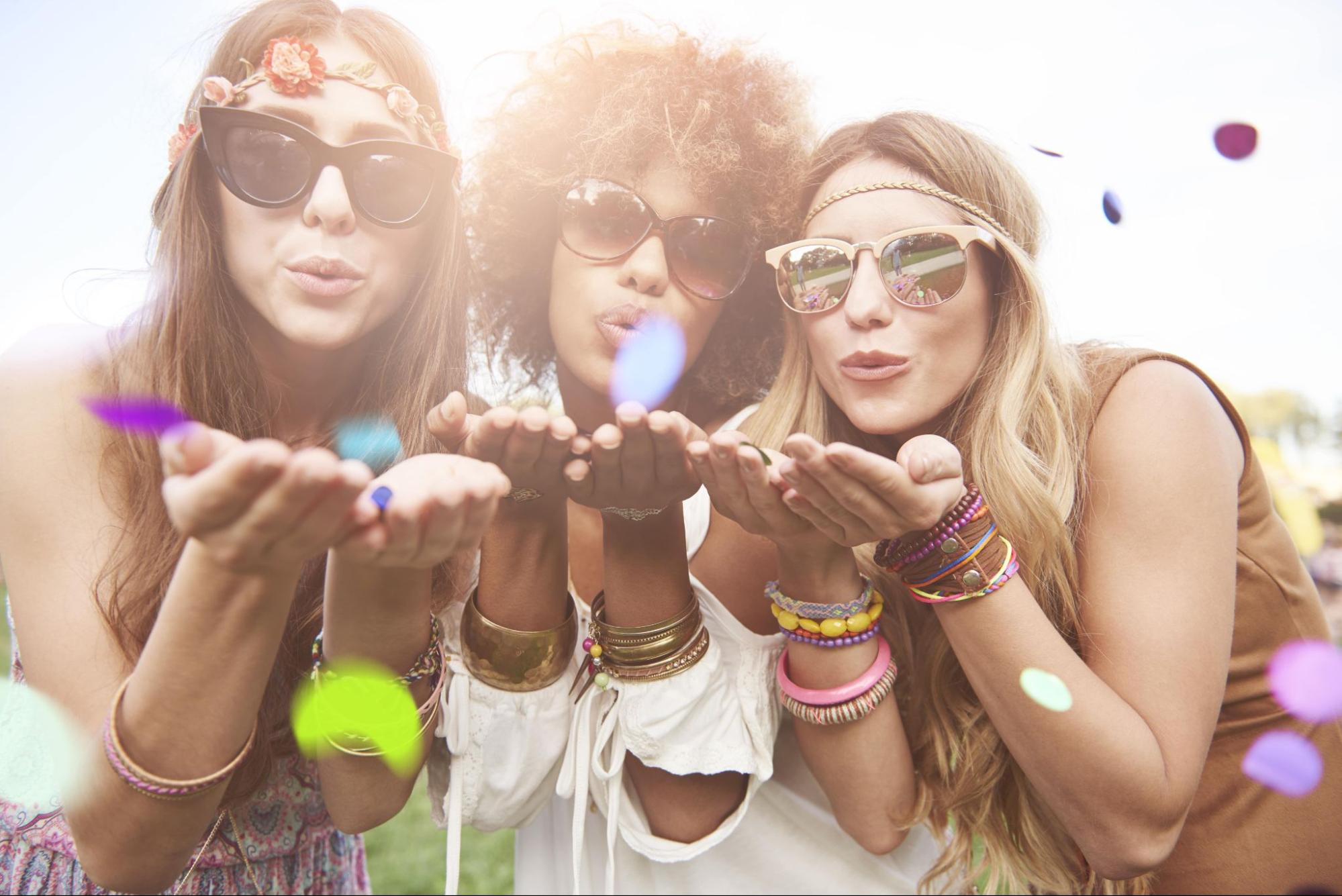 Three women in sunglasses and colorful layered bracelets blew confetti towards camera