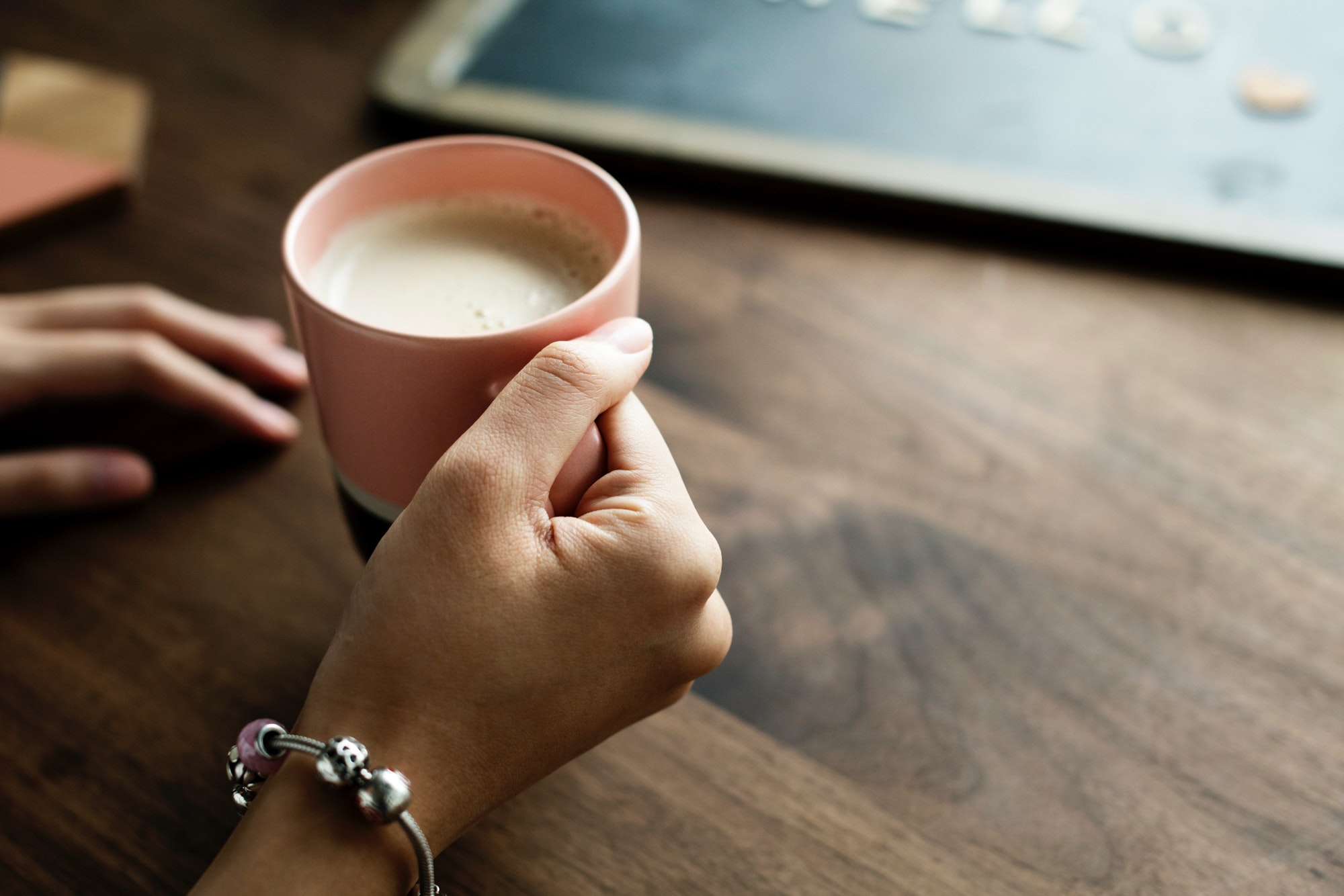 A woman holding a pink coffee mug sat at a wooden desk, her silver bracelet visible.