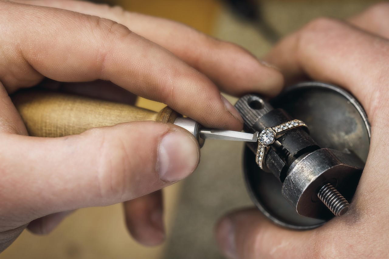 A jeweler's hands intricately detail a diamond ring held in a metal clamp.