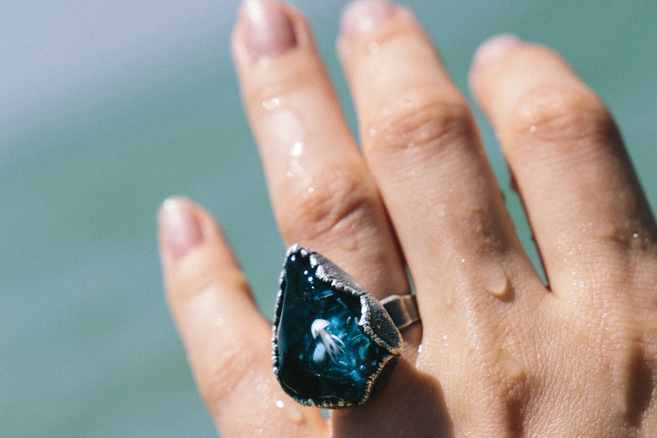 Close-up of a hand with water droplets, wearing a blue teardrop-shaped ring against a blurred background.