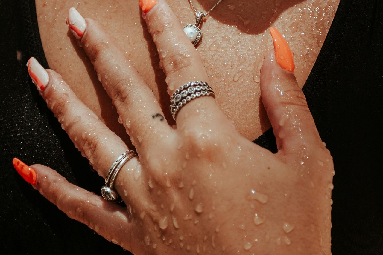 Close-up of a hand with wet skin on a chest, adorned with rings and vibrant orange and white nails.