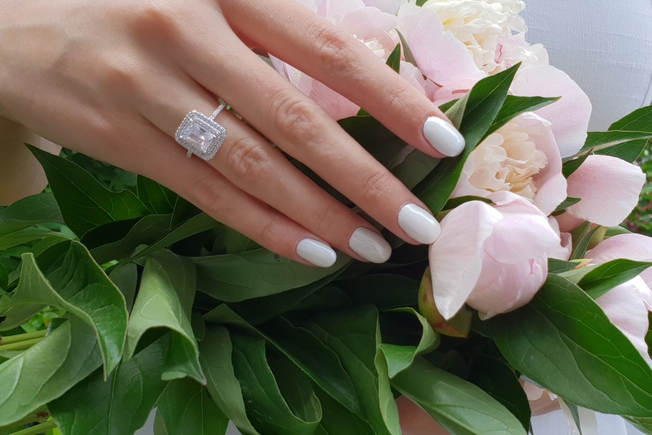 A graceful hand with white manicured nails and a sparkling emerald cut diamond ring rests on pink peonies and green leaves.