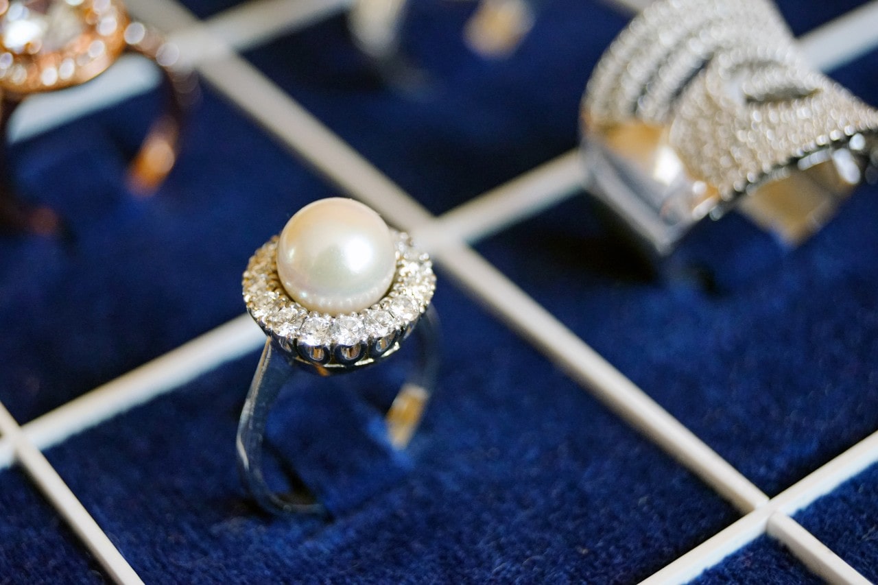 A pearl ring with a diamond-encrusted band displayed on a dark blue velvet grid, alongside other sparkling, elegant jewelry pieces.