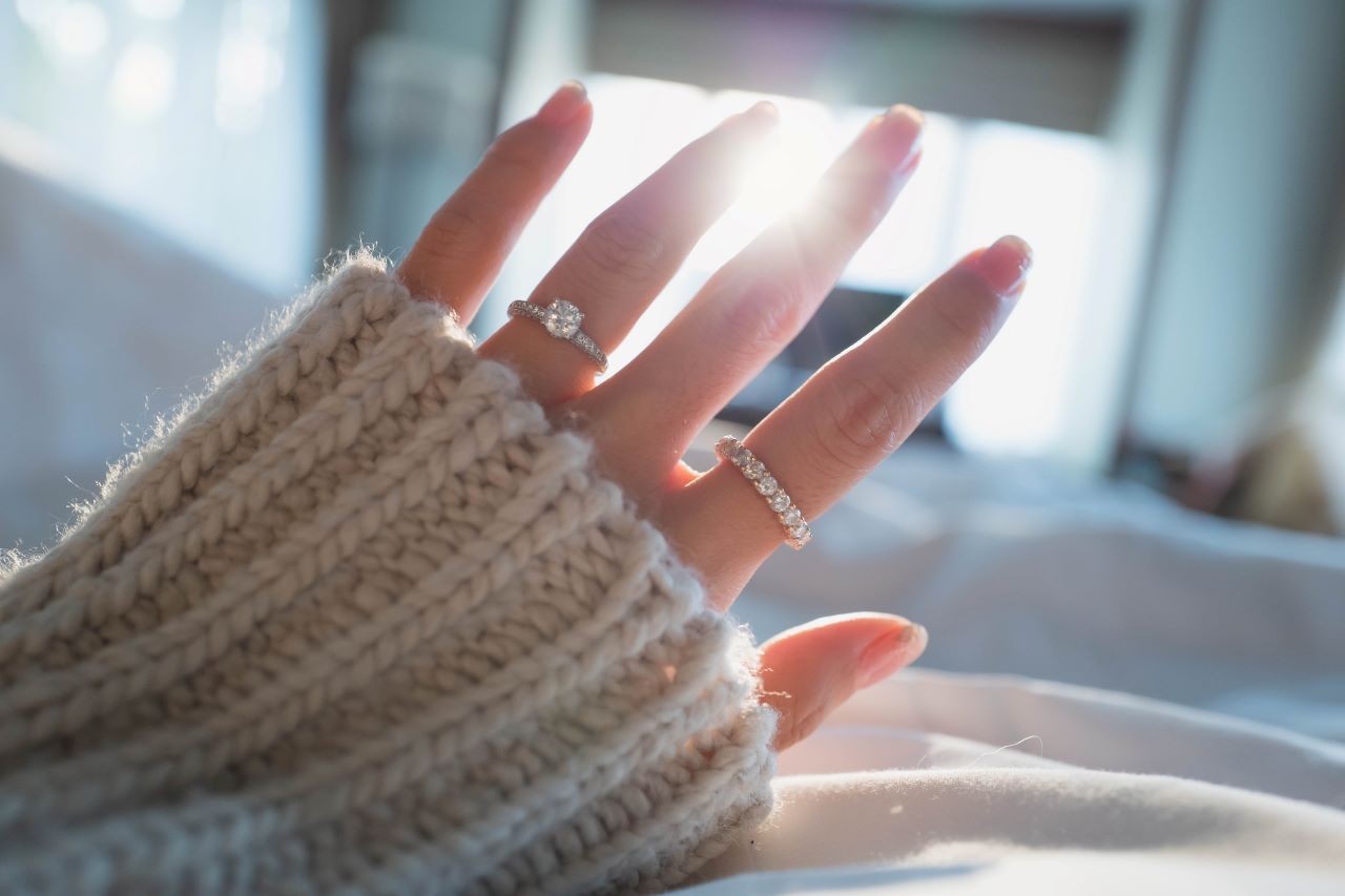 A hand adorned with sparkling diamond rings is raised against a sunlit room.