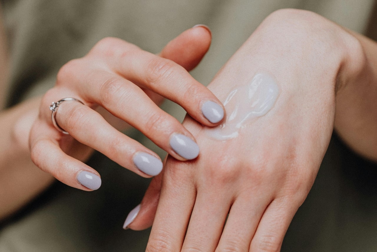 A close-up of a woman&rsquo;s hands with a solitaire ring and light grey nails, applying cream onto the back of her hand.