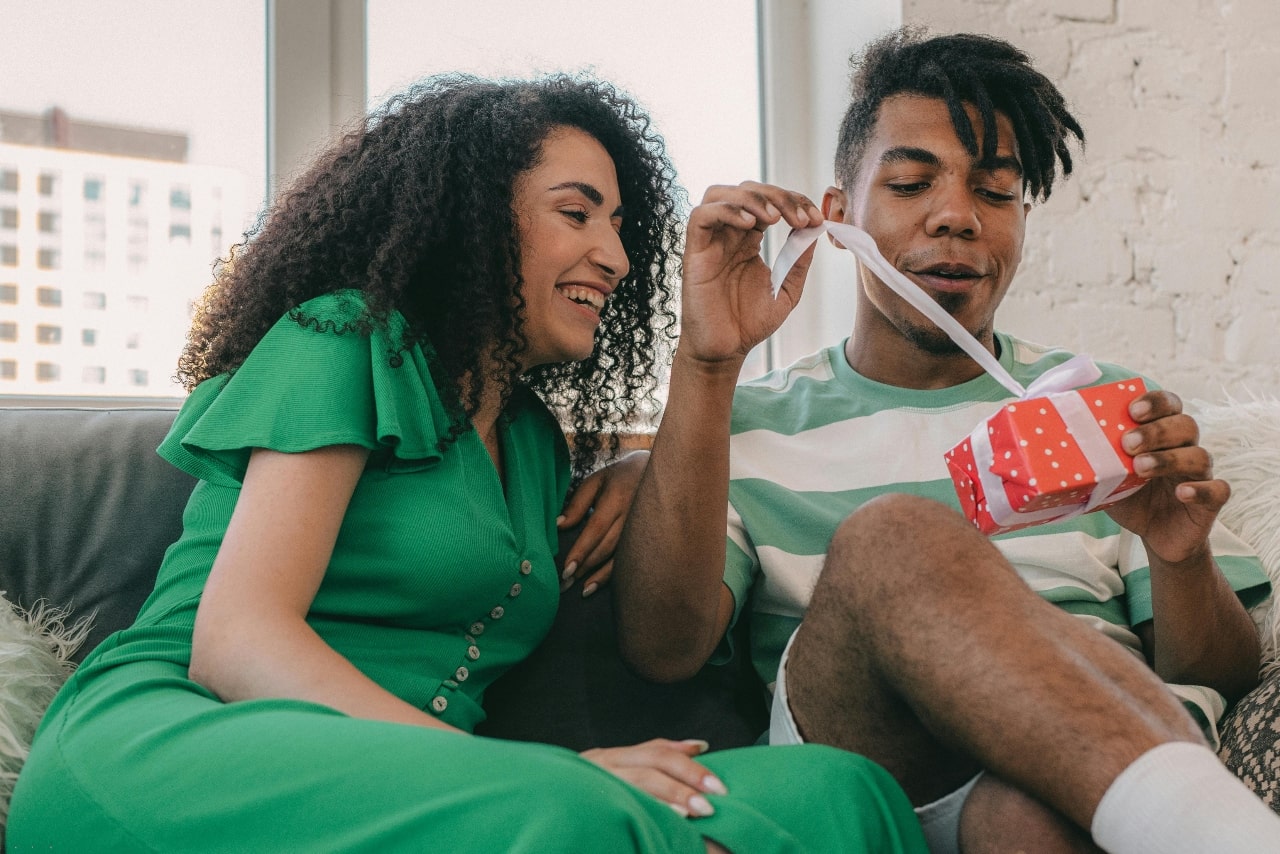 A view of a young man opening a small red gift box with a white ribbon while sitting beside a smiling woman in a green dress.