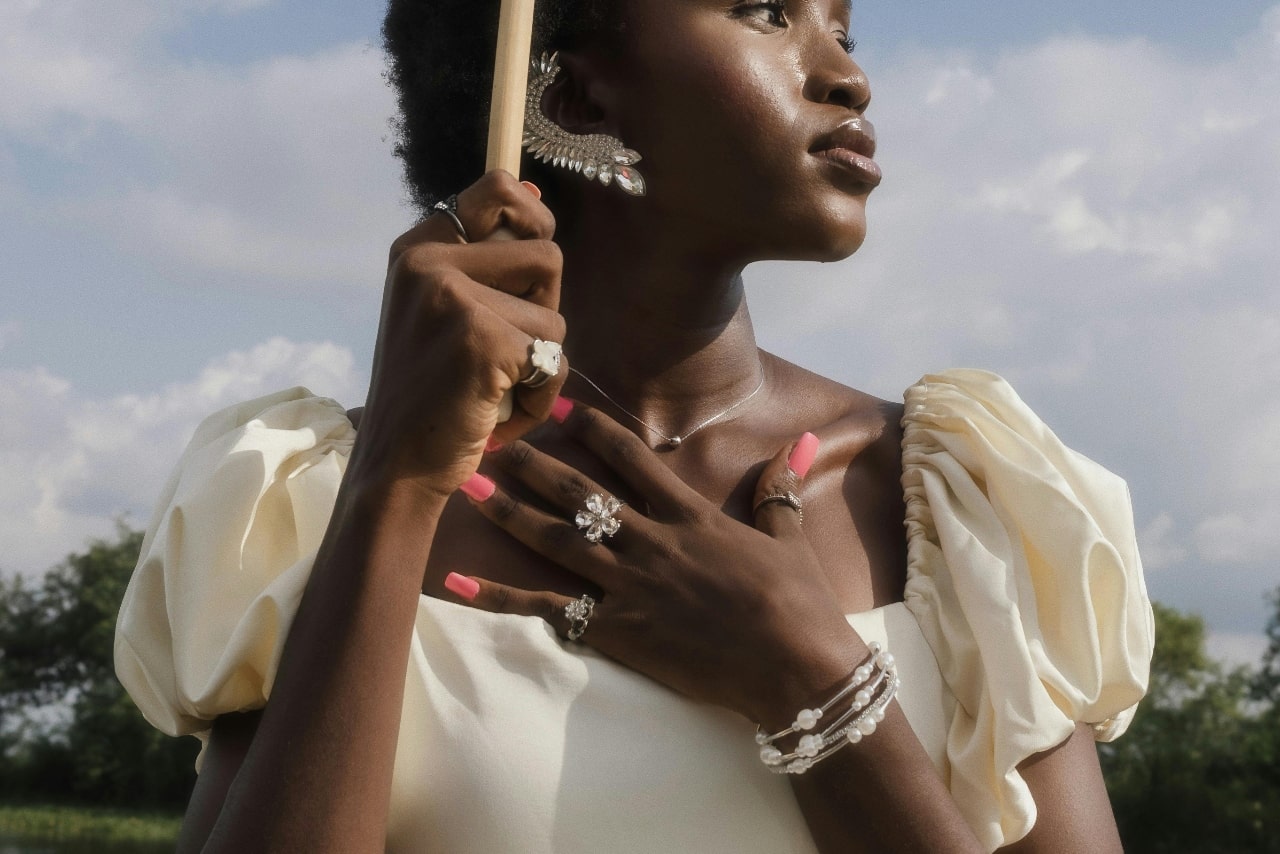 A woman holding her hand to her chest in an open field, with emphasis on the eyecatching jewelry on her hands, neck, and ear.