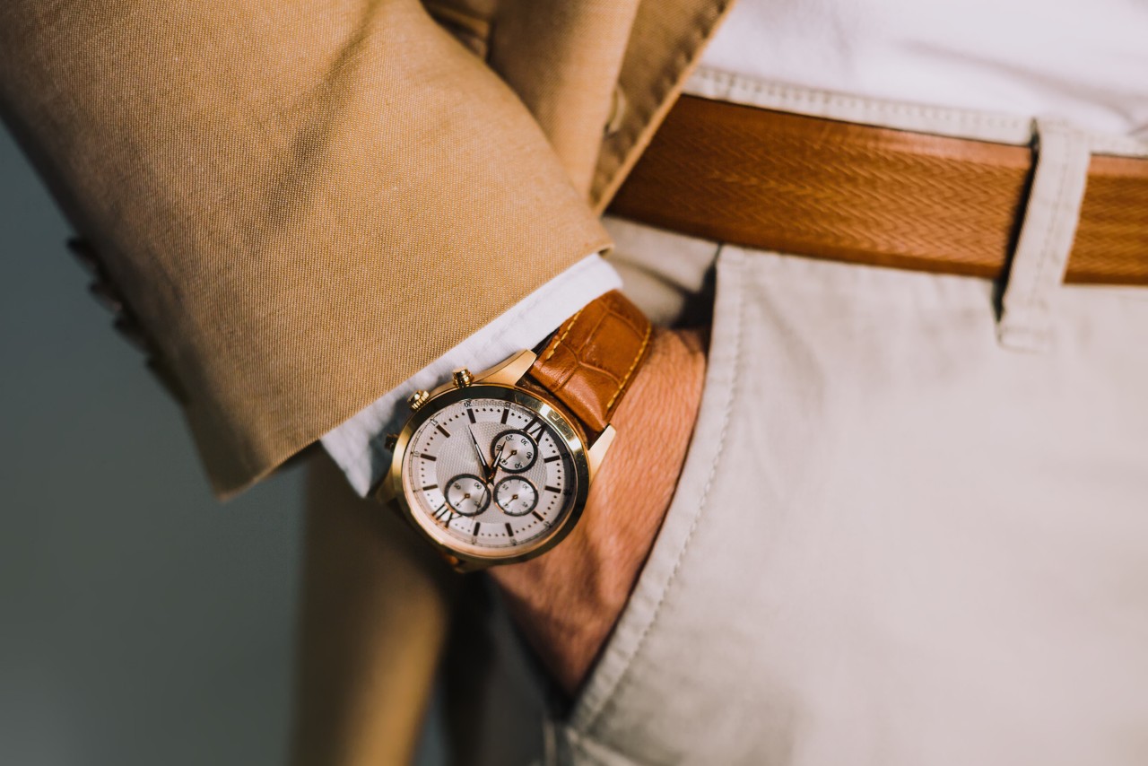 A close up of a man with his hands in the pockets of beige trousers, showcasing a wristwatch with brown leather and crisp white dial.