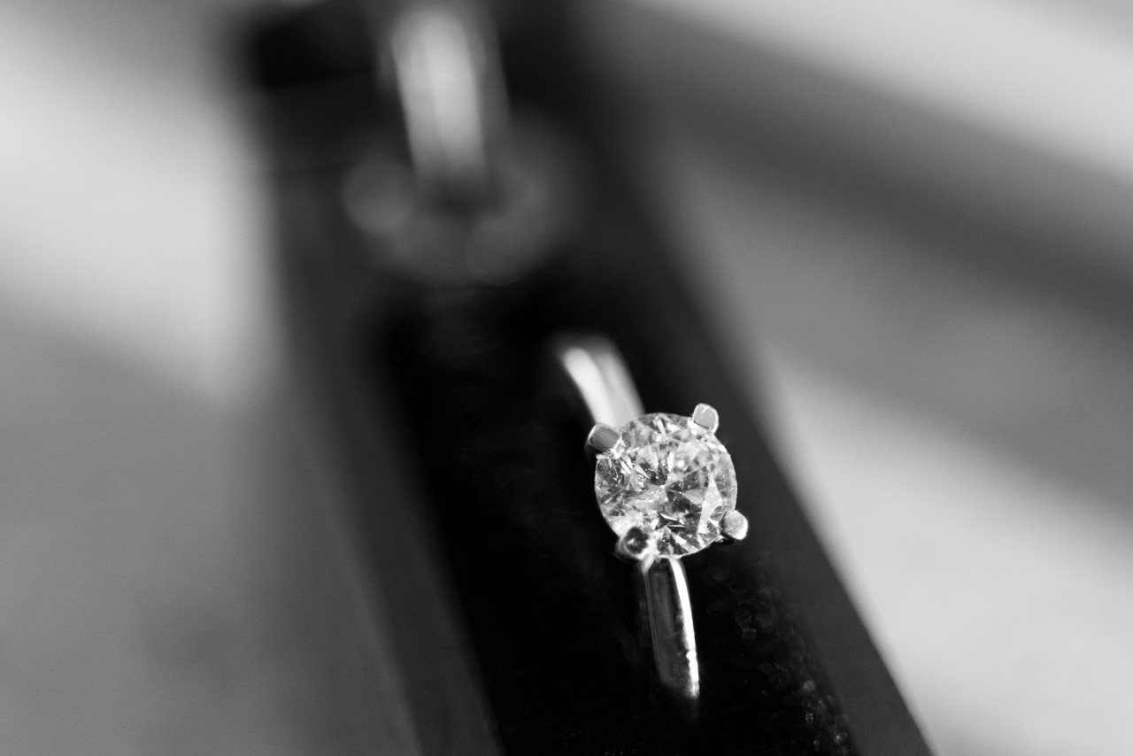 A black and white image of a solitaire engagement ring displayed upright inside a ring holder.