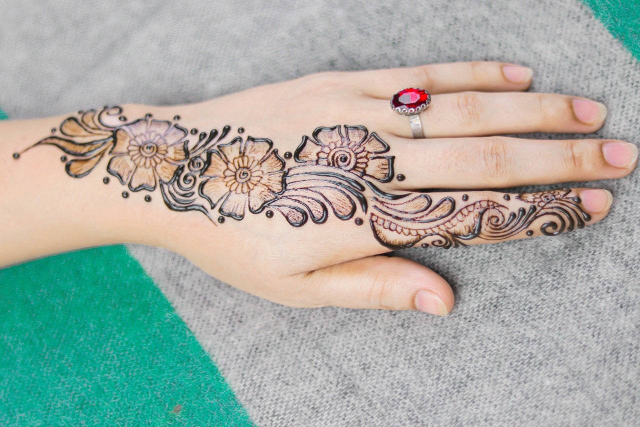 A person&rsquo;s hand adorned with henna flowers and a bright ruby and silver ring.