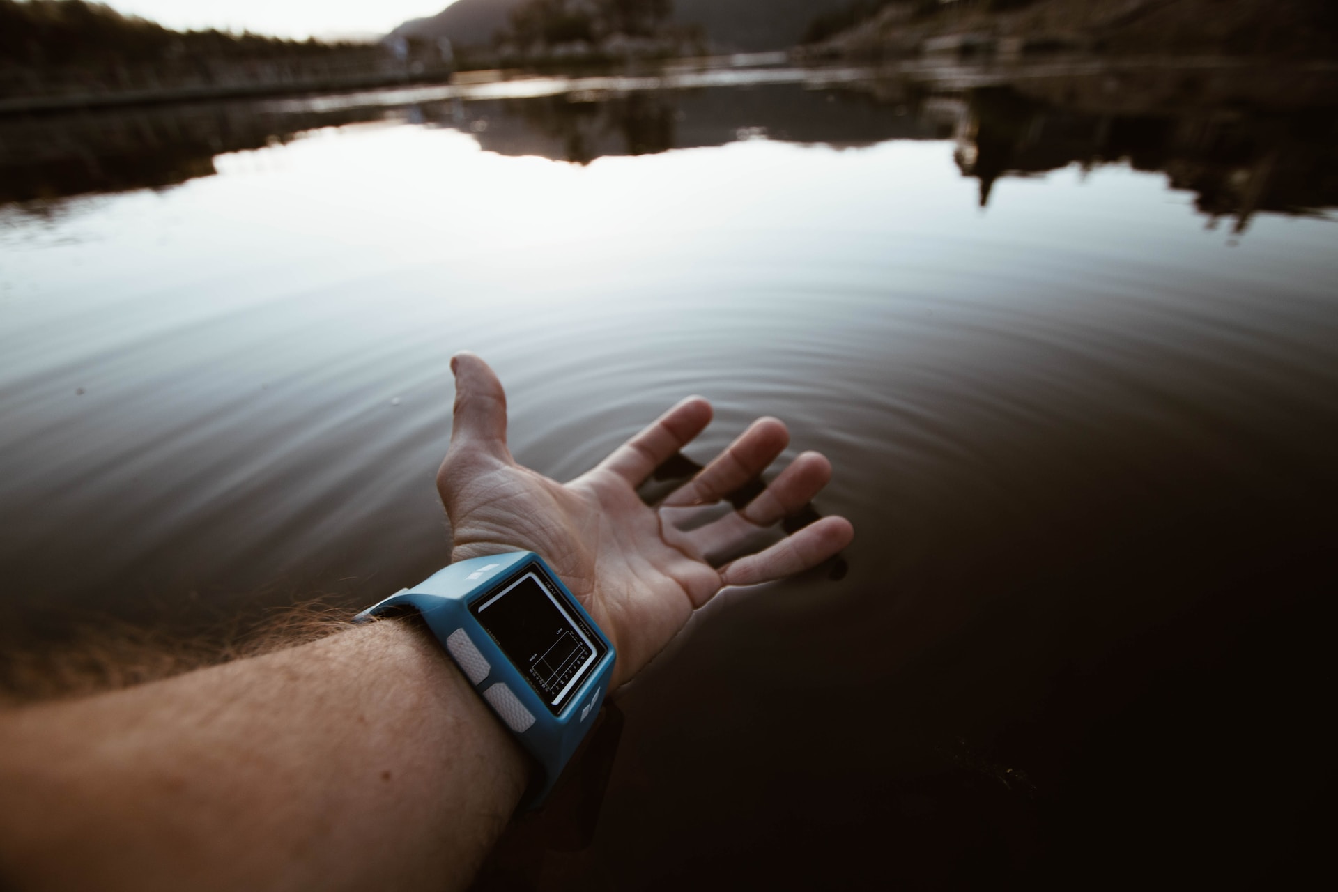 A hand reaches over calm water, creating gentle ripples. The wrist wears a blue smartwatch.