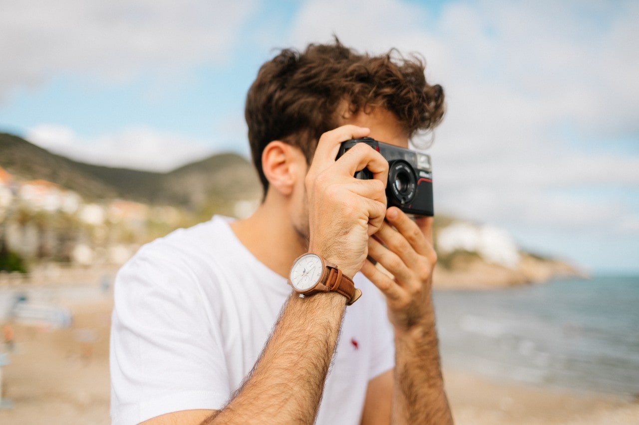 A person in a white shirt, wearing an elegant wrist watch holds a camera to their face, focusing intently.