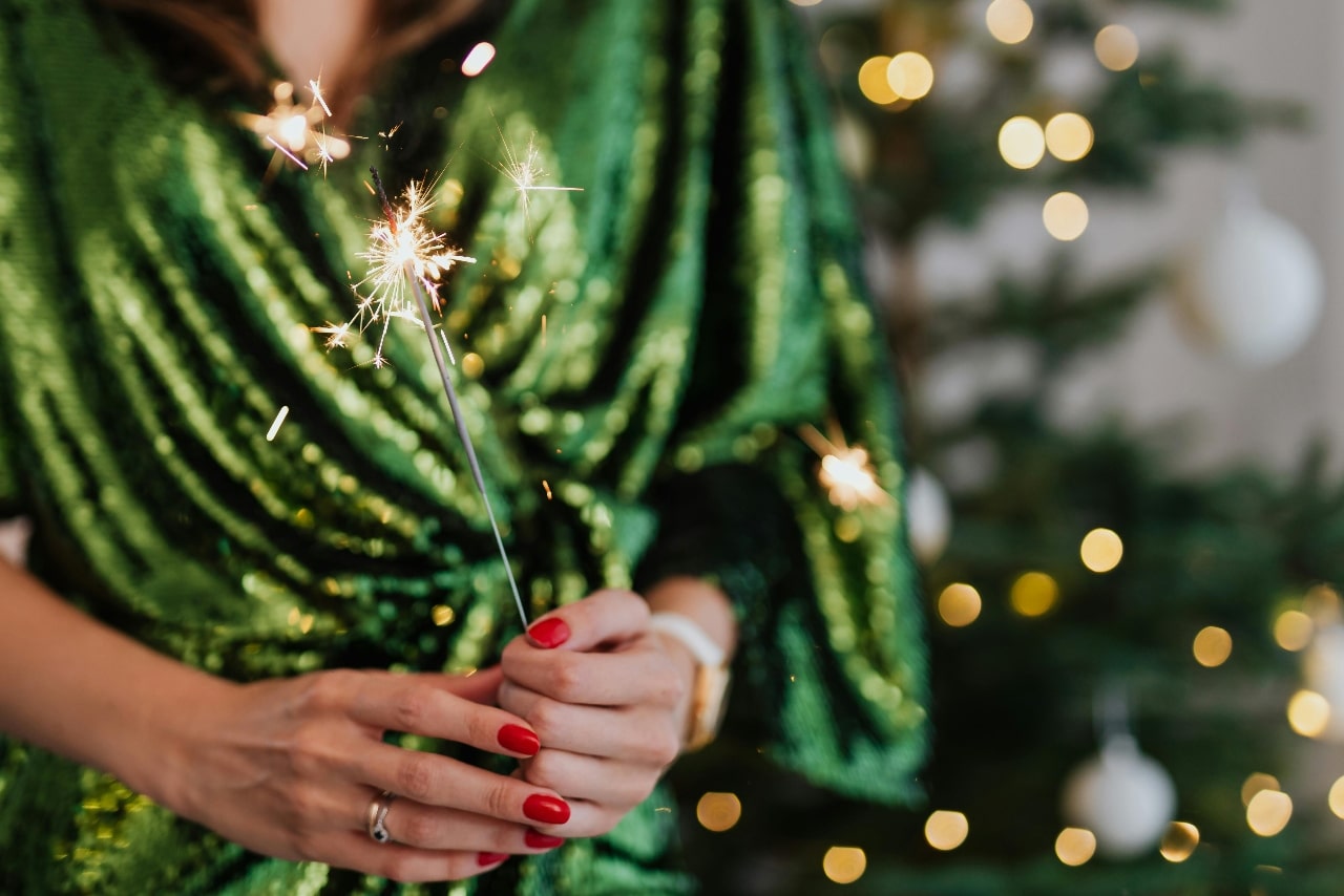 Woman in a shimmering green dress holding a lit sparkler in front of a blurred Christmas tree
