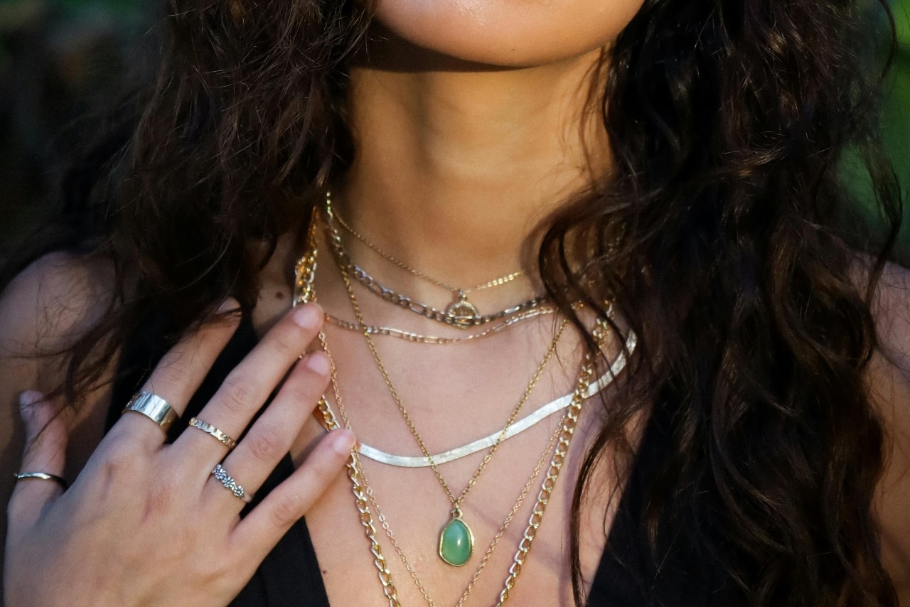 Woman wearing multiple layered gold necklaces of varying lengths, including a green teardrop pendant, paired with gold rings and a black top.