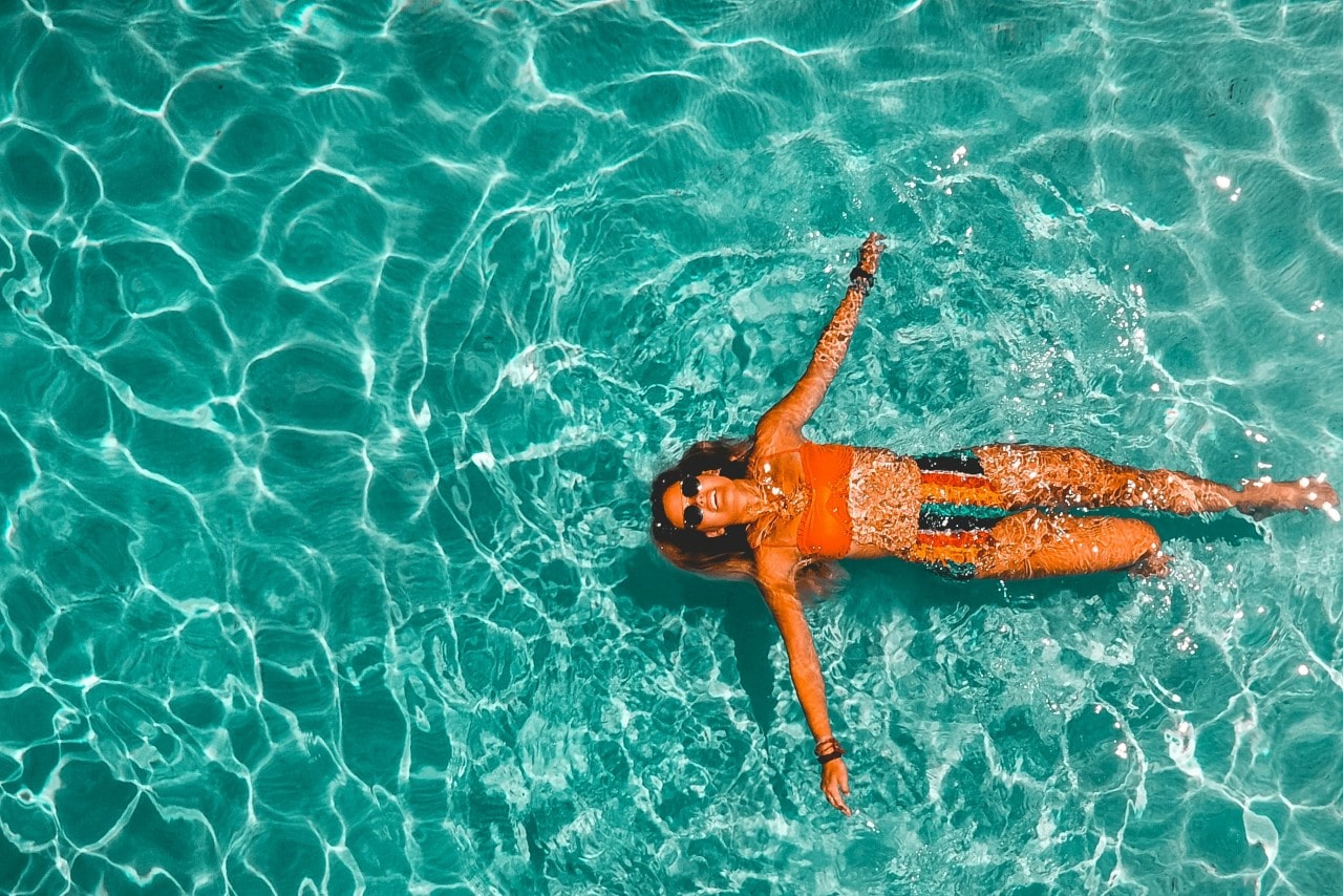 A woman in an orange swimsuit swims on top of crystal blue sunlit water.